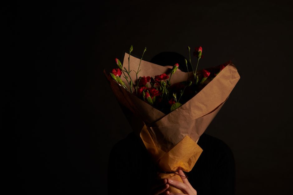 A person holding a floral bouquet wrapped in brown kraft paper, featuring vibrant red and pink flowers with green stems and leaves. The bouquet is positioned in front of a dark background, highlightin