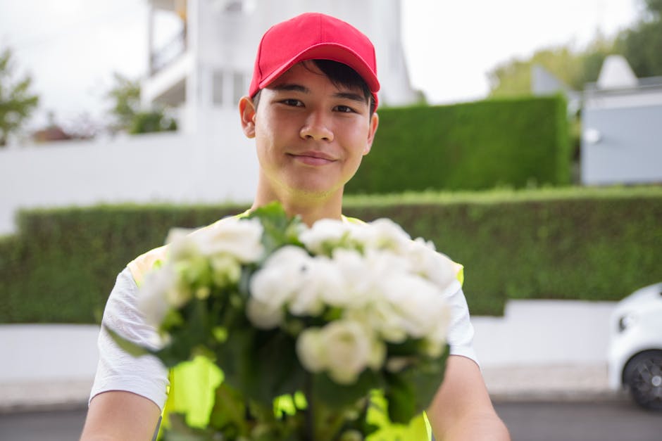 A male flower delivery courier wearing a red baseball cap and high-visibility vest is standing outdoors on a residential street, smiling as he presents a bouquet of fresh white roses and green foliage