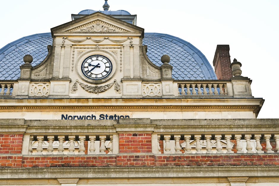 A close-up view of the ornate facade of Norwich Station, featuring a large round clock with black Roman numerals and hands set against a white background, surrounded by intricate stone carvings and de