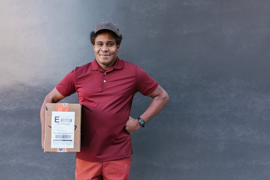A delivery person with dark skin and short, curly hair, wearing a maroon polo shirt, matching shorts, a gray cap, and a black wristwatch, holding a small cardboard package with a shipping label in the