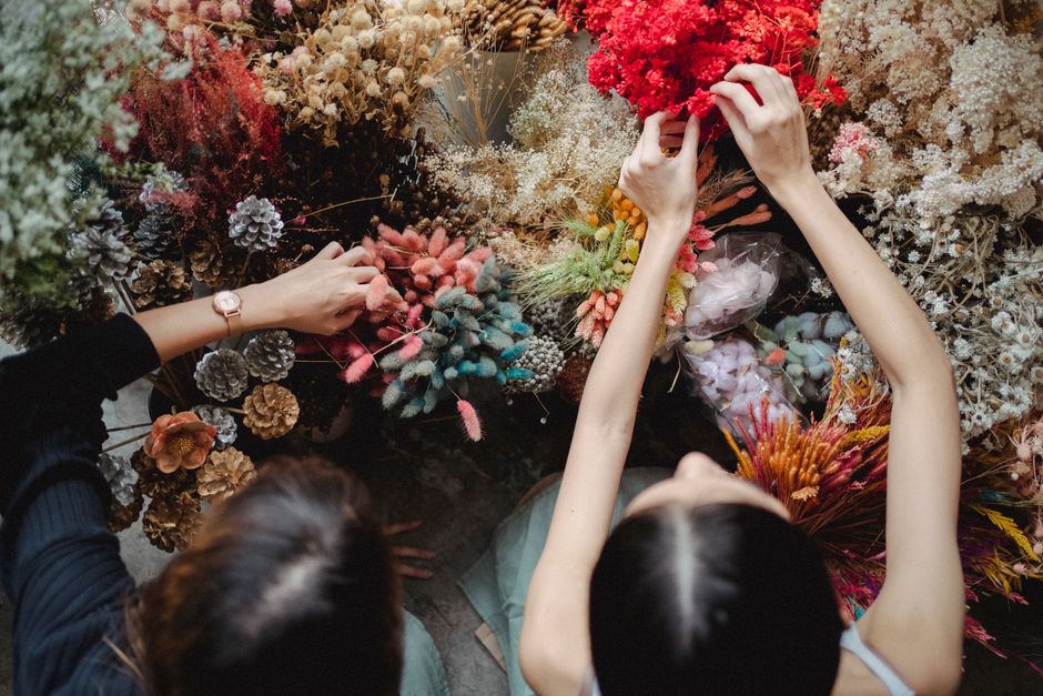 Two florists working at a table filled with a diverse array of fresh flowers, including pink, white, purple, and red blooms, as well as dried floral elements like pine cones, all arranged in various s