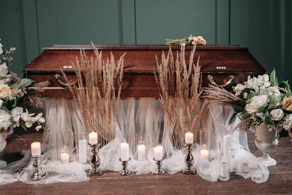 A wooden funerary casket with a polished surface is placed on a dark wooden floor, surrounded by floral arrangements and candles. Tall, feathery dried wheat stalks are positioned at either end of the 