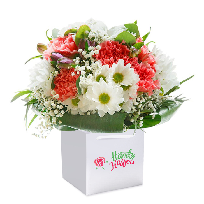 Elegantly arranged by a skilled Mottingham florist, this radiant bouquet is presented upright in a minimalist white square gift bag, making a vibrant statement against a spotless, white backdrop. The bouquet's composition is a testament to local craftsmanship, featuring plush coral and blush carnations with distinctive ruffled petals, evocative of spring gardens along Court Farm Road. White daisies, with their crisp petals and vivid lime-green cores, punctuate the arrangement, injecting a joyful brightness. Wispy sprigs of baby's breath meander through the mass of blooms, providing an ethereal, cloud-like lightness. Nestled on the left, subtle hints of unopened, purple-blushed buds lend depth, suggesting growth and the promise of future blossoms. The entire floral ensemble is encircled by robust, glossy green foliage, arranged in sweeping arcs to form a sumptuous, rounded silhouette with a sense of fullness and completeness. The clean white ribbon handles atop the bag ensure practical elegance, while the neutral background accentuates every colour, petal, and leaf detail. More than just a gift, this arrangement embodies Mottingham's sense of community-ideal for celebrations, milestone anniversaries, or simply sending a heartfelt thought to someone special in the local area.