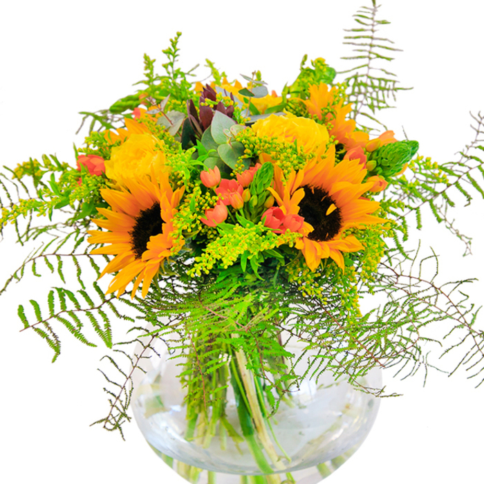 The first impression of this Mottingham bouquet is one of joyful sunlight and the comfort of home. Large, radiant sunflowers-each with a deep brown heart and a halo of vibrant yellow-orange petals-anchor this extraordinary arrangement, reminiscent of the cheerful wildflower fields around Mottingham's green spaces. Between these bright blooms nestle classic yellow garden roses, their tightly curled petals unfolding like golden promises. Among them, brilliant clusters of red Hypericum berries glisten, catching the light and providing a playful pop of color and texture. The entire sense of abundance is enhanced by an array of lush leaves: delicate, cascading ferns introduce gentle movement and a touch of woodland tranquility, while sprays of subtle goldenrod and green flower spikes add fresh detail and depth. Wisps of blue-green eucalyptus foliage peek through for a gentle, aromatic lift. Encased in a sparkling, rounded glass vase, the bouquet reveals its verdant stems bathed in clear water-a symbol of freshness and lasting vitality. Designed with both artistry and heart by experienced Mottingham florists, this arrangement is an ideal choice for birthdays, celebrations, or simply bringing spirited color into homes along Mottingham Road, seamlessly echoing the warmth and unity of the local community.